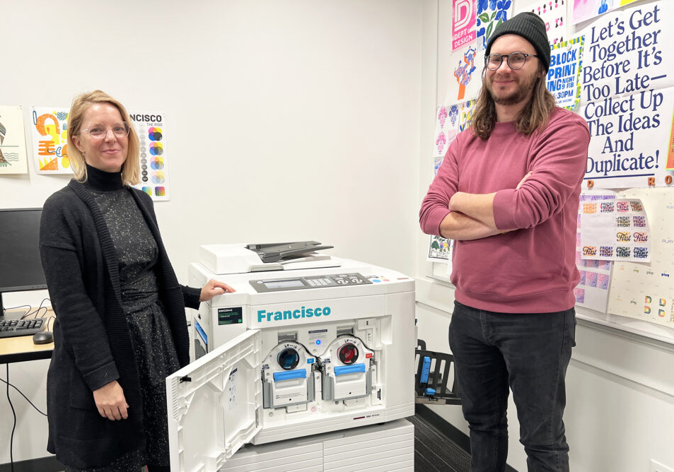 Two design faculty stand near a Risograph, in winter 2024, that was added to the 4D Lab.