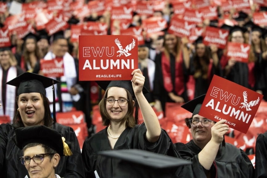 A crowd of college graduates in gowns in mortar boards hold signs that say "EWU alumni."
