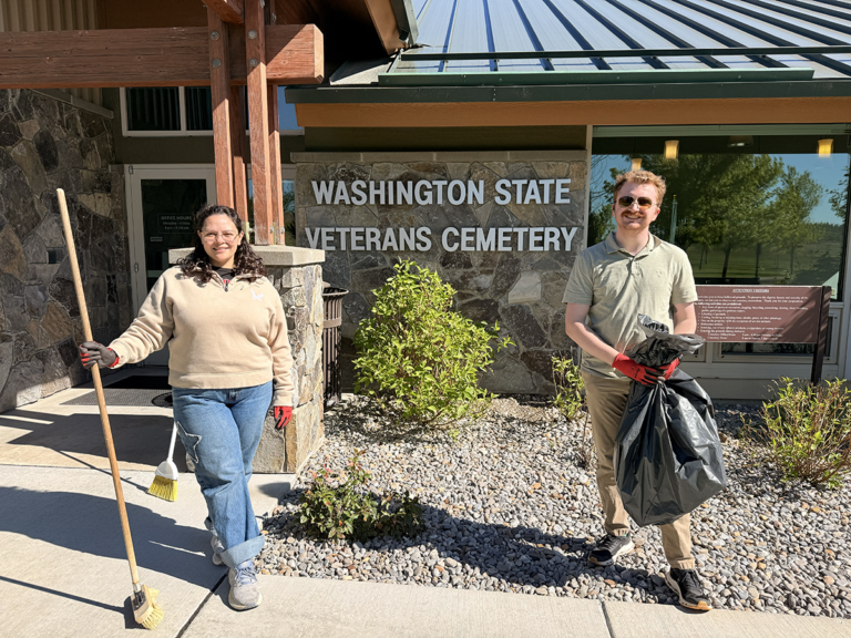 Two student volunteers with a broom and trash bag pose with the Washington State Veterans Cemetery sign behind them.